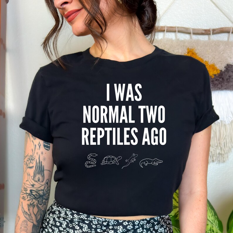 May include: Black t-shirt with white text that reads "I was normal two reptiles ago". The text is surrounded by small white illustrations of a snake, turtle, lizard, and alligator.