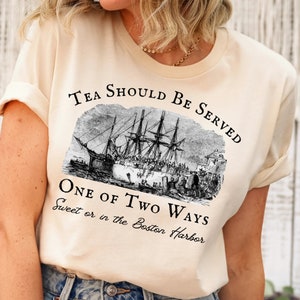 May include: A beige t-shirt with a black graphic print of a ship and the text "Tea Should Be Served One Of Two Ways Sweet or in the Boston Harbor".