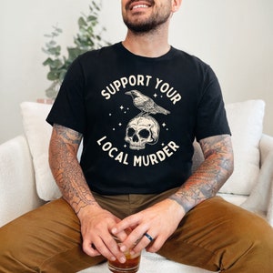 May include: A black t-shirt with a graphic design of a crow perched on a skull. The text "Support Your Local Murder" is printed in a white font around the graphic.