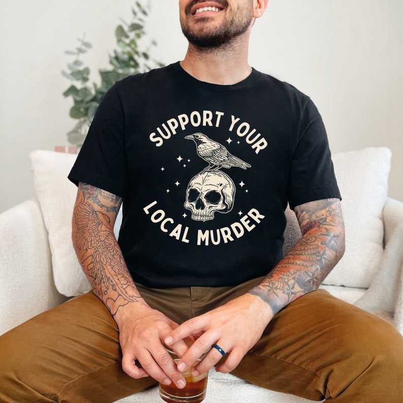 May include: A black t-shirt with a graphic design of a crow perched on a skull. The text "Support Your Local Murder" is printed in a white font around the graphic.