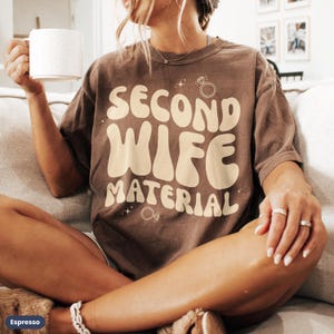 May include: A brown t-shirt with the text "Second Wife Material" in a retro font. The text is in a light beige color. There are two small diamond ring illustrations on the shirt.