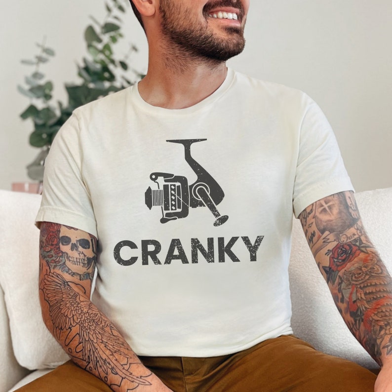 May include: A white t-shirt with a black fishing reel graphic and the text "Cranky" printed on the front.