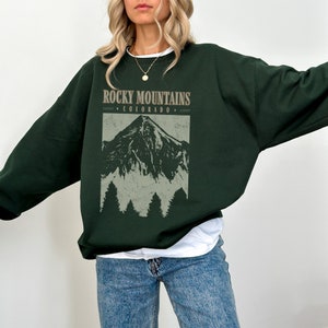 May include: A dark green sweatshirt with a vintage-style graphic of the Rocky Mountains and the text "Rocky Mountains - Colorado -" printed on the front.
