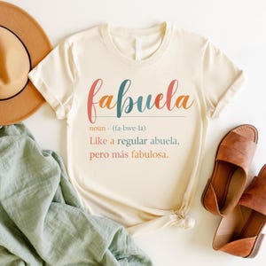 Puede incluir: Una camiseta blanca con la palabra "fabuela" escrita en letras de colores. La definici&oacute;n de la palabra es "Como una abuela normal, pero m&aacute;s fabulosa."