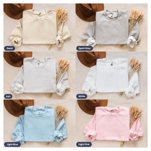 May include: Six different color sweatshirts laid flat on a light brown surface. The sweatshirts are all the same style, with long sleeves and a crew neck. The colors are sand, sport grey, ash, white, light blue, and light pink.