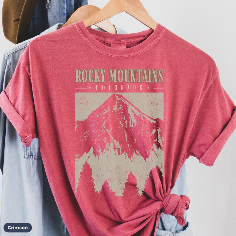 May include: Crimson red t-shirt with a vintage-style graphic of a mountain range and the text "Rocky Mountains - Colorado -" printed on the front.
