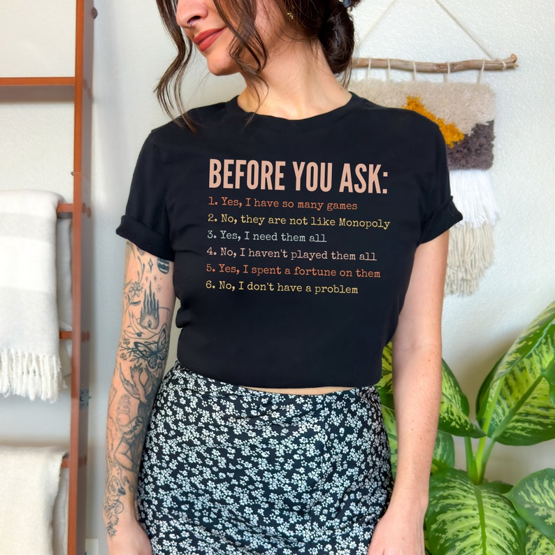 K&ouml;nnte beinhalten: Ein schwarzes T-Shirt mit dem Text "BEFORE YOU ASK:" und einer Liste von Fragen und Antworten in einem Farbverlauf von Rosa, Orange und Gelb. Der Text lautet: "1. Ja, ich habe so viele Spiele. 2. Nein, sie sind nicht wie Monopoly. 3. Ja, ich brauche sie alle. 4. Nein, ich habe nicht alle gespielt. 5. Ja, ich habe ein Verm&ouml;gen daf&uuml;r ausgegeben. 6. Nein, ich habe kein Problem."