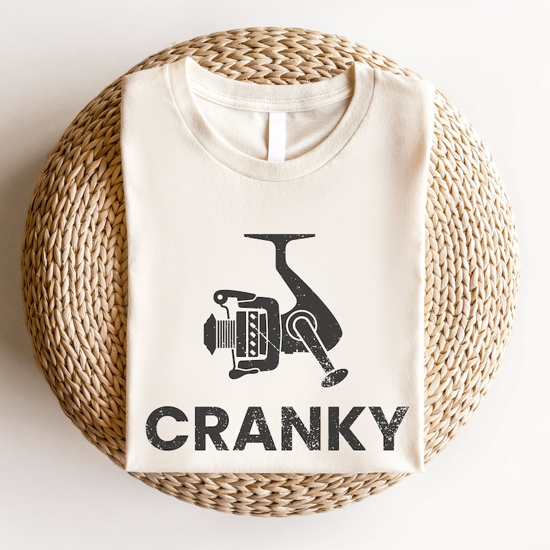 May include: A beige t-shirt with a black graphic of a fishing reel and the word "CRANKY" printed on it.