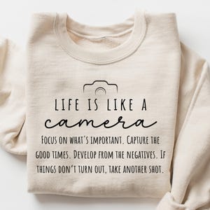 May include: A beige sweatshirt with a black graphic of a camera and the text "LIFE IS LIKE A camera FOCUS ON WHAT'S IMPORTANT. CAPTURE THE GOOD TIMES. DEVELOP FROM THE NEGATIVES. IF THINGS DON'T TURN OUT, TAKE ANOTHER SHOT."