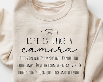 May include: A beige sweatshirt with a black graphic of a camera and the text "LIFE IS LIKE A camera FOCUS ON WHAT'S IMPORTANT. CAPTURE THE GOOD TIMES. DEVELOP FROM THE NEGATIVES. IF THINGS DON'T TURN OUT, TAKE ANOTHER SHOT."