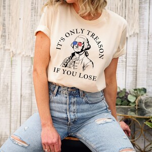 May include: A beige t-shirt featuring a graphic of George Washington wearing sunglasses with the American flag on them. The text on the shirt reads "It's only treason if you lose."