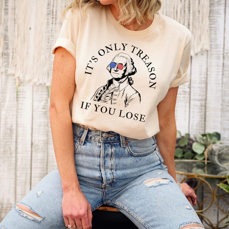 May include: A beige t-shirt featuring a graphic of George Washington wearing sunglasses with the American flag on them. The text on the shirt reads "It's only treason if you lose."