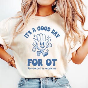 May include: A white t-shirt with blue text that reads "It's a good day for OT. Movement is medicine."  A blue cartoon hand with a smiling face is walking and holding a star.