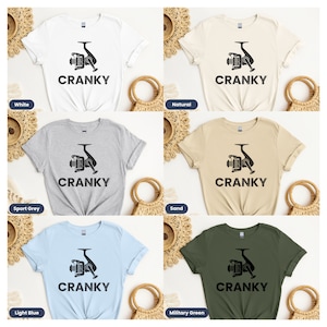 May include: Six different color t-shirts with a black and white graphic of a fishing reel and the word "CRANKY" printed on the front. The colors are white, sport grey, light blue, natural, sand, and military green.