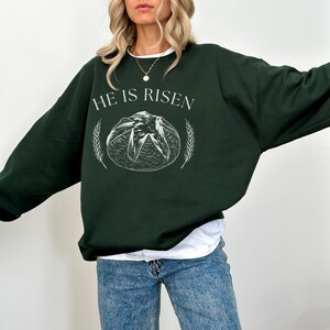May include: Dark green sweatshirt with the text "HE IS RISEN" in white. The design includes a white illustration of a loaf of bread and wheat stalks. The sweatshirt is a long-sleeved, crew neck style.
