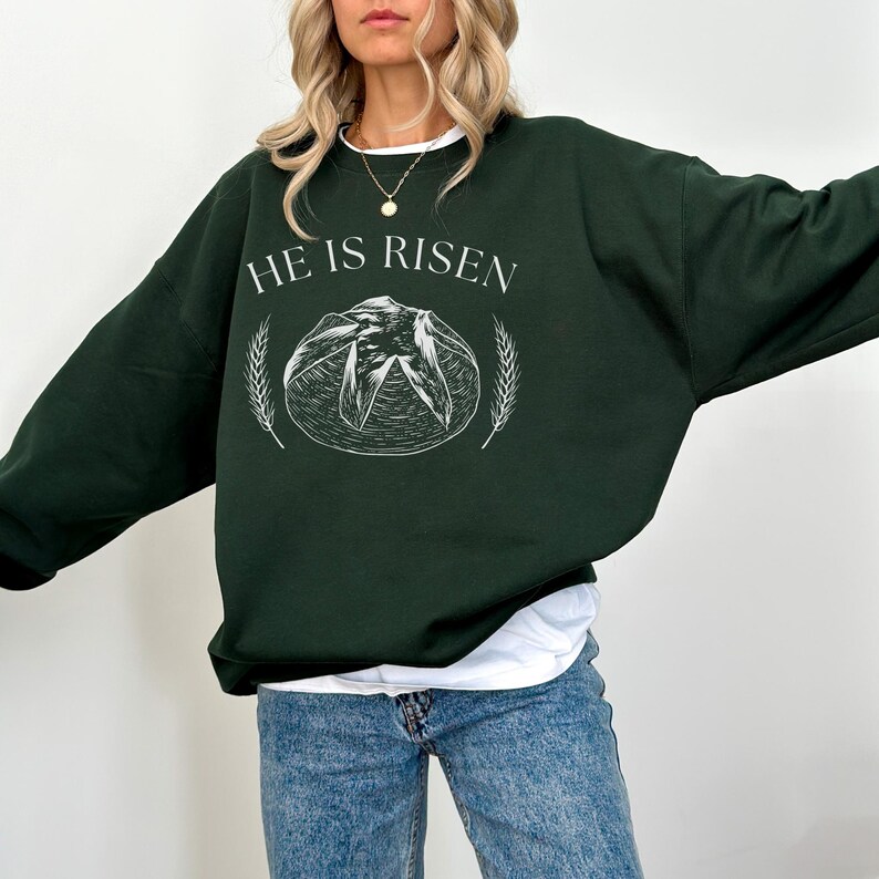 May include: Dark green sweatshirt with the text "HE IS RISEN" in white. The design includes a white illustration of a loaf of bread and wheat stalks. The sweatshirt is a long-sleeved, crew neck style.