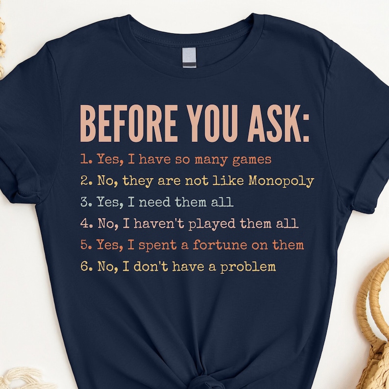 K&ouml;nnte beinhalten: Ein marineblaues T-Shirt mit dem Text "BEFORE YOU ASK:" und einer Liste von sechs humorvollen Aussagen &uuml;ber Brettspiele. Der Text ist in einer bunten Schriftart.