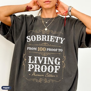 May include: A dark gray t-shirt with a vintage-style design that reads "Sobriety From 100 Proof to Living Proof Premium Edition".