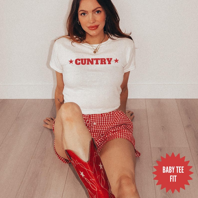 May include: White cropped t-shirt with red "COUNTRY" text and red stars. The model is wearing the t-shirt with red and white gingham shorts and red cowboy boots. The text "BABY TEE FIT" is in a red starburst.
