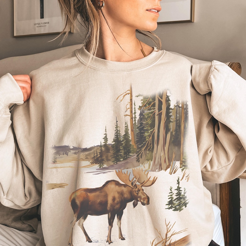 May include: A beige sweatshirt with a moose and forest scene graphic print. The moose is brown and has large antlers. The forest scene is a watercolor style with green trees and a blue sky.