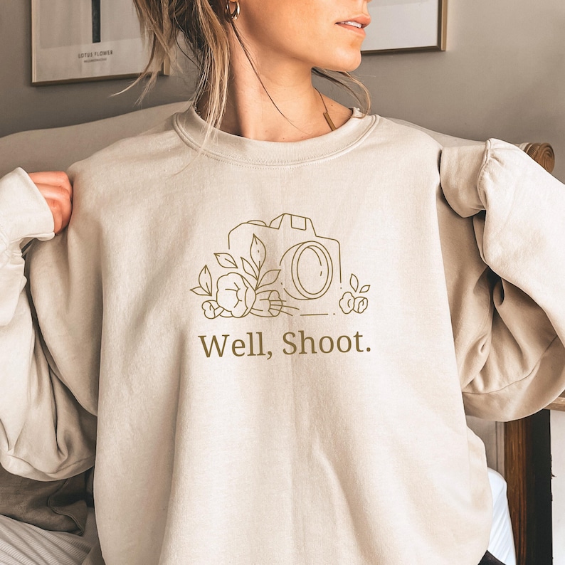 May include: A beige sweatshirt with a graphic of a camera and flowers. The text "Well, Shoot." is printed below the graphic.