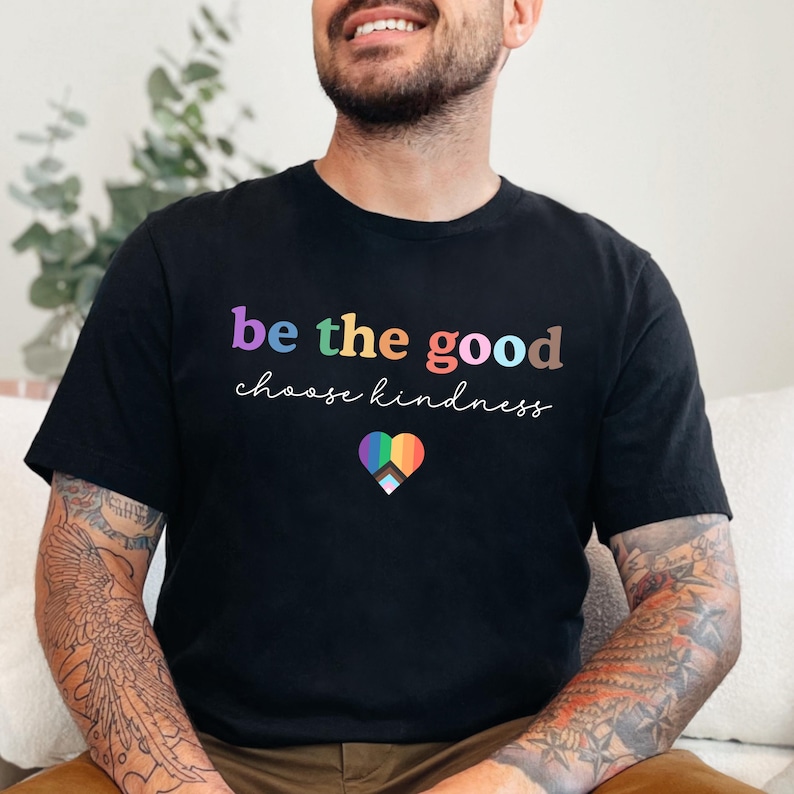 Może przedstawiać: Czarny T-shirt z napisem "be the good choose kindness" w kolorach tęczy i tęczowym sercem z czarno-białym symbolem pokoju w środku.