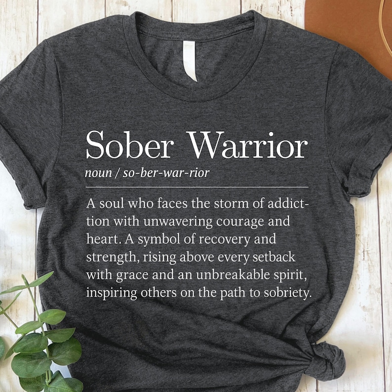 May include: A dark gray t-shirt with white text that reads "Sober Warrior" and a definition of the term. The definition describes a person who faces challenges with courage and strength.