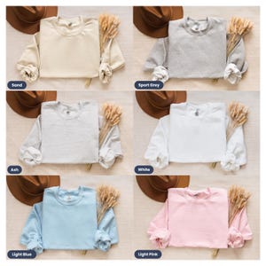 May include: Six different color sweatshirts laid flat on a light brown surface. The colors are sand, sport grey, ash, white, light blue, and light pink.