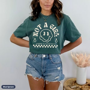 May include: A green t-shirt with a vintage-style smiley face graphic and the text "Not a Girl" in a circle. The text "Gender is a construct" is printed below the graphic. The shirt is worn by a person with long brown hair and is paired with denim shorts.