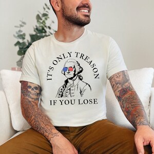 May include: A white t-shirt featuring a black and white illustration of George Washington wearing sunglasses with the American flag on them. The text on the shirt reads "It's only treason if you lose."