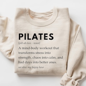Pu&ograve; includere: Una felpa beige con la parola "PILATES" in testo nero, seguita da una definizione: "[pill-ah-tees - noun] Un allenamento mente-corpo che trasforma lo stress in forza, il caos in calma e le giornate brutte in giornate migliori. vedi anche: il mio happy hour"