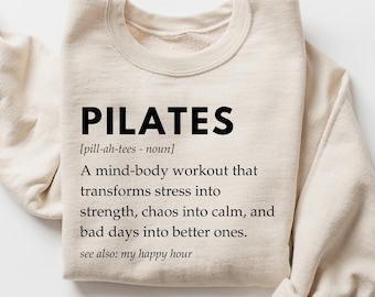 May include: A beige sweatshirt with the word "PILATES" in black text, followed by a definition: "[pill-ah-tees - noun] A mind-body workout that transforms stress into strength, chaos into calm, and bad days into better ones. see also: my happy hour"