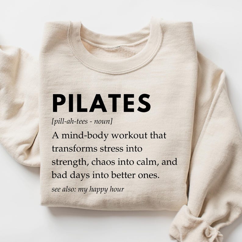 Pu&ograve; includere: Una felpa beige con la parola "PILATES" in testo nero, seguita da una definizione: "[pill-ah-tees - noun] Un allenamento mente-corpo che trasforma lo stress in forza, il caos in calma e le giornate brutte in giornate migliori. vedi anche: il mio happy hour"