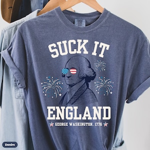 May include: A blue t-shirt with a white graphic of George Washington wearing sunglasses with the American flag design. The text "SUCK IT ENGLAND" is above the image and "GEORGE WASHINGTON. 1776" is below the image.