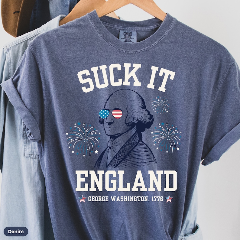 May include: A blue t-shirt with a white graphic of George Washington wearing sunglasses with the American flag design. The text "SUCK IT ENGLAND" is above the image and "GEORGE WASHINGTON. 1776" is below the image.