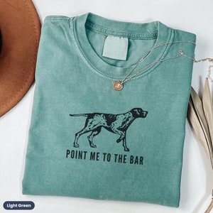 May include: Light green t-shirt with a black and white illustration of a dog pointing. The text "POINT ME TO THE BAR" is printed below the dog.