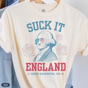 May include: A cream-colored t-shirt with a blue and red graphic of George Washington wearing sunglasses with the American flag on them. The graphic also includes fireworks and the text "SUCK IT ENGLAND GEORGE WASHINGTON, 1776".