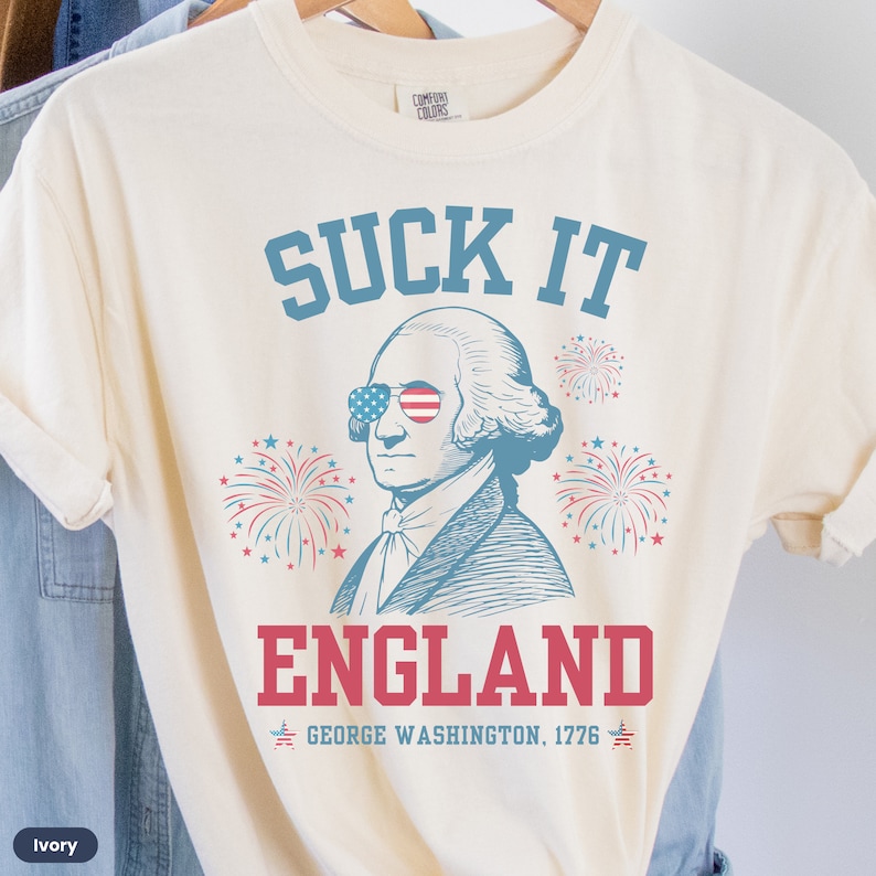 May include: A cream-colored t-shirt with a blue and red graphic of George Washington wearing sunglasses with the American flag on them. The graphic also includes fireworks and the text "SUCK IT ENGLAND GEORGE WASHINGTON, 1776".