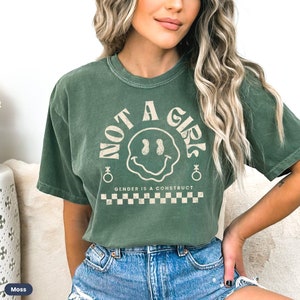 May include: A green t-shirt with a vintage-style graphic that says "Not a Girl" with a smiley face and the text "Gender is a construct" below it. The shirt has a checkered pattern at the bottom.