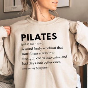 Pu&ograve; includere: Una felpa beige con la parola "PILATES" in testo nero. Il testo sotto definisce il Pilates come un allenamento mente-corpo che trasforma lo stress in forza, il caos in calma e le giornate brutte in giornate migliori. Il testo dice anche "vedi anche: il mio happy hour".
