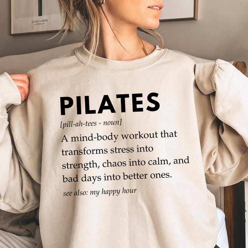 Pu&ograve; includere: Una felpa beige con la parola "PILATES" in testo nero. Il testo sotto definisce il Pilates come un allenamento mente-corpo che trasforma lo stress in forza, il caos in calma e le giornate brutte in giornate migliori. Il testo dice anche "vedi anche: il mio happy hour".