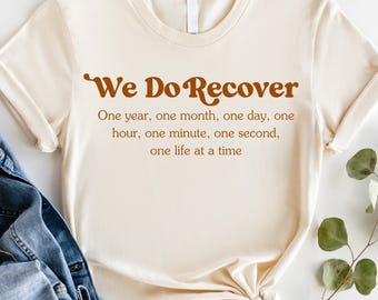 May include: A beige t-shirt with the text 'We Do Recover' in brown lettering. The text continues with 'One year, one month, one day, one hour, one minute, one second, one life at a time'.