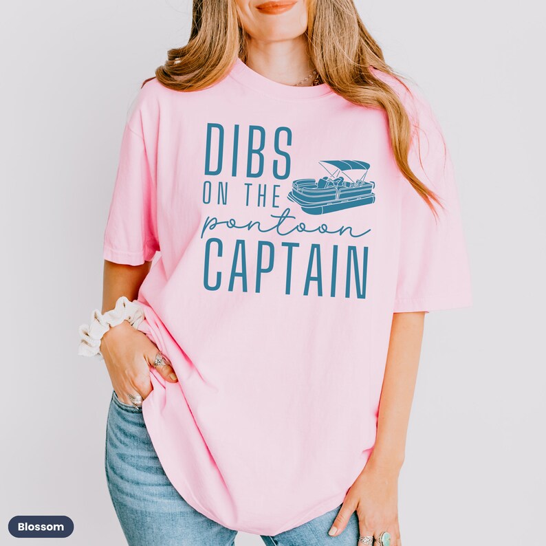 May include: Pink t-shirt with the text "DIBS ON THE pontoon CAPTAIN" in blue lettering. A blue illustration of a pontoon boat is above the text.