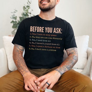K&ouml;nnte beinhalten: Ein schwarzes T-Shirt mit dem Text "BEFORE YOU ASK:" und einer Liste von Fragen und Antworten in humorvollem Ton. Der Text ist in einem wei&szlig;en, orangefarbenen und rosafarbenen Farbschema gehalten.