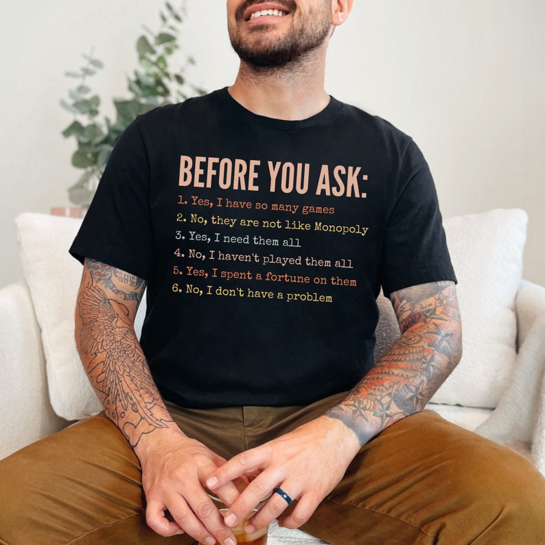 K&ouml;nnte beinhalten: Ein schwarzes T-Shirt mit dem Text "BEFORE YOU ASK:" und einer Liste von Fragen und Antworten in humorvollem Ton. Der Text ist in einem wei&szlig;en, orangefarbenen und rosafarbenen Farbschema gehalten.
