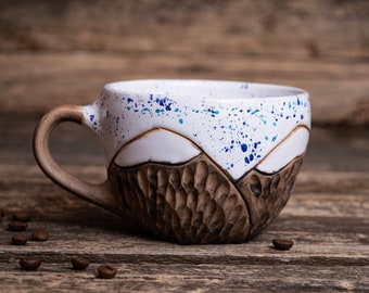 Może przedstawiać: Ręcznie robiony ceramiczny kubek do kawy z unikalnym górskim wzorem. Kubek ma białe wnętrze z niebieskimi plamami, brązowy uchwyt i teksturowany, rzeźbiony brązowy wygląd zewnętrzny. Ziarna kawy są rozrzucone.