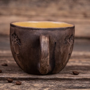May include: A brown ceramic mug with a yellow interior and a tree design. The mug is sitting on a wooden surface.