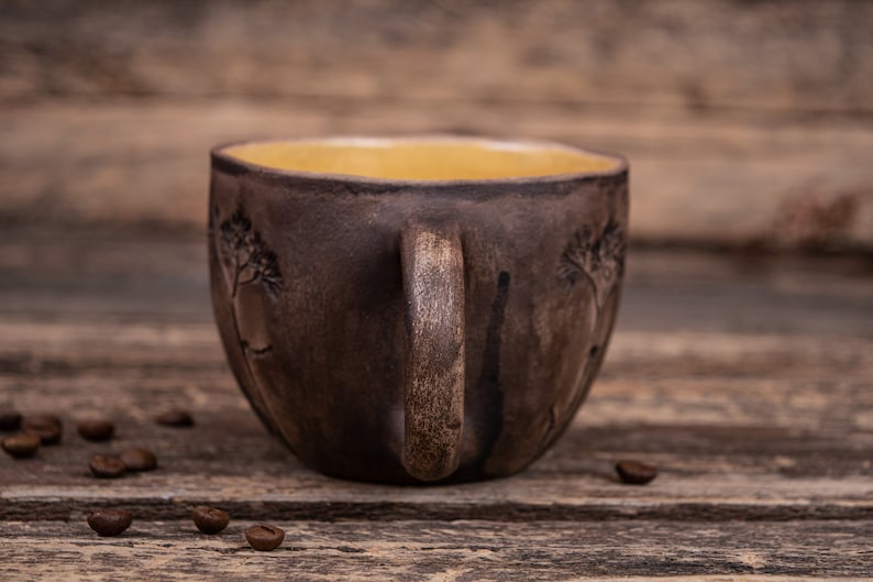 May include: A brown ceramic mug with a yellow interior and a tree design. The mug is sitting on a wooden surface.