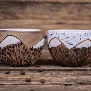Może przedstawiać: Dwa ceramiczne kubki z wzorem g&oacute;r. Jeden kubek jest brązowy z białymi g&oacute;rami, a drugi jest biały z niebieskimi plamkami i białymi g&oacute;rami.