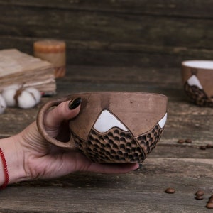 Może przedstawiać: Brązowy kubek ceramiczny z białym wzorem g&oacute;r. Kubek jest fakturowany i ma uchwyt. Kubek jest trzymany przez rękę z czerwoną bransoletką.
